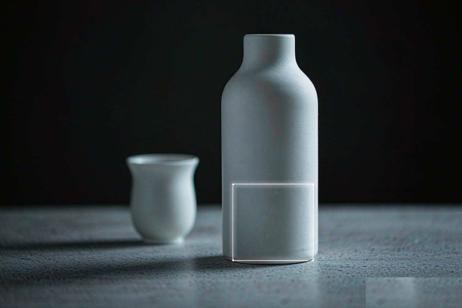 A white ceramic bottle and cup on a grey stone surface, with a thin glowing outline marking one small edited region while the rest stays unchanged
