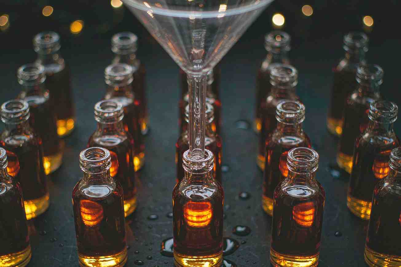 Muchas botellas de vidrio pequeñas e idénticas llenándose desde un único embudo de vidrio situado encima sobre una superficie oscura