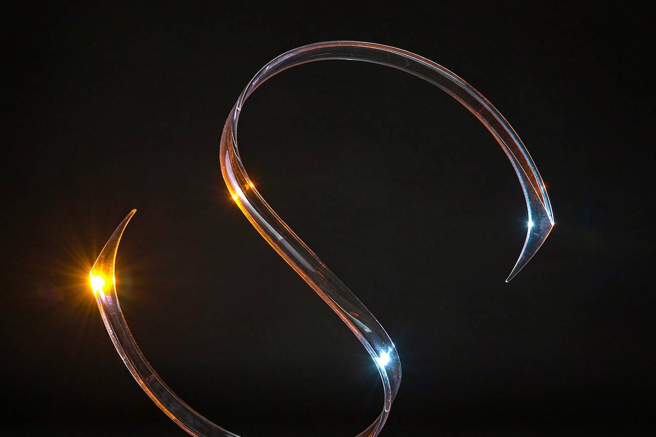 A single ribbon of clear glass curving through darkness, lit warm and cool