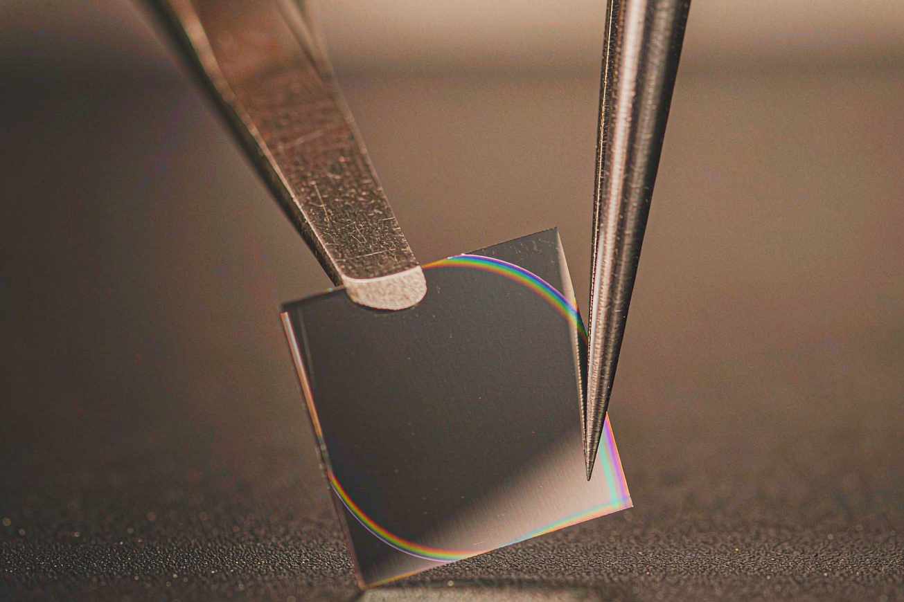 Tweezers holding a square silicon die with iridescent reflections, suspended over a warm-toned lab bench