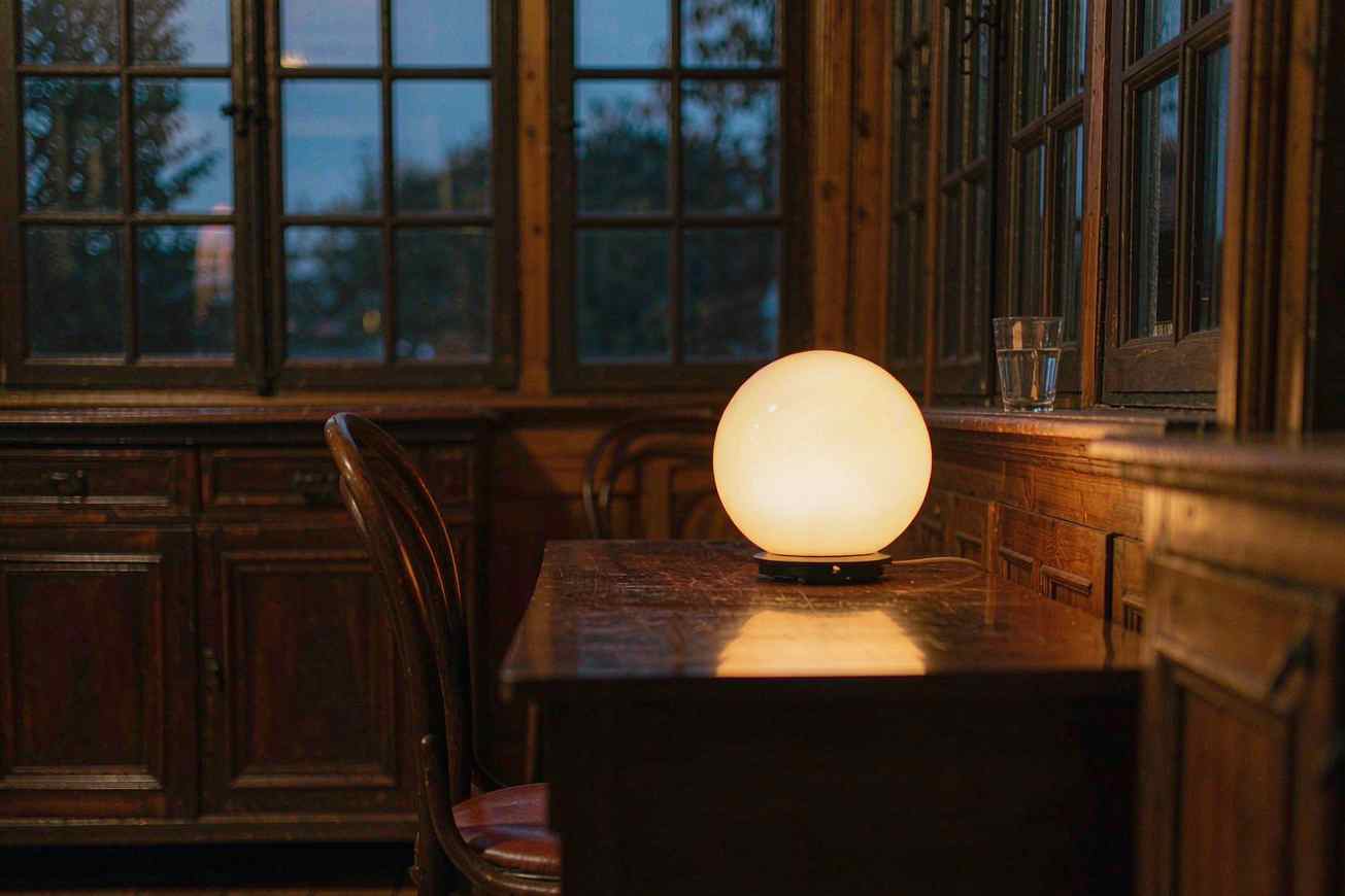 Une lampe ronde en verre allumée sur une table en bois foncé près d'une grande fenêtre au crépuscule