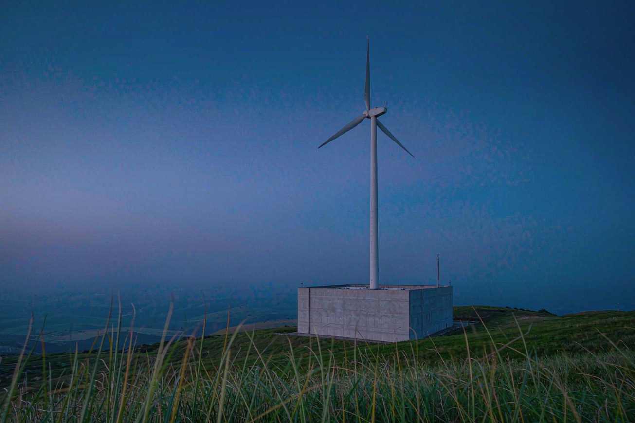 A plain concrete substation beside a tall white wind turbine on a green hill at blue hour