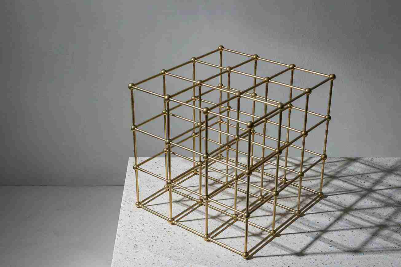 An open geometric wireframe cube of thin brass rods resting on a pale concrete plinth