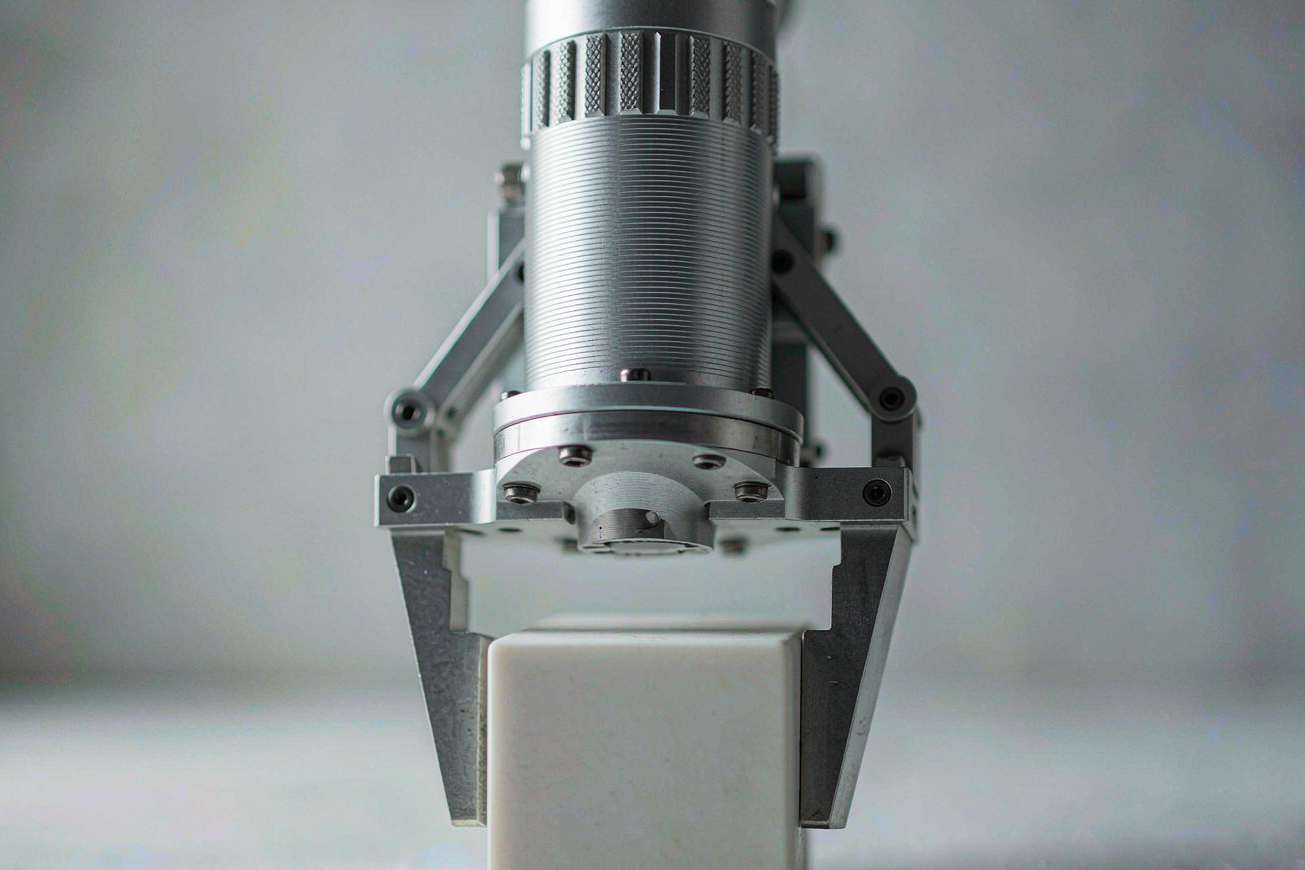 A brushed aluminium robotic gripper approaching a plain white ceramic cube on a pale grey surface