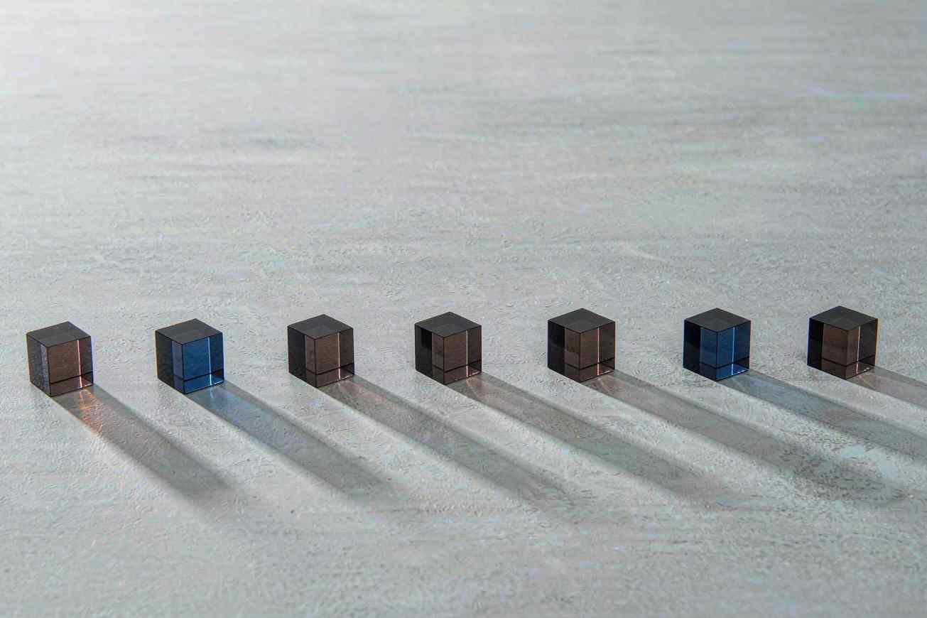 Seven small translucent colored cubes arranged in a precise arc on a pale concrete surface