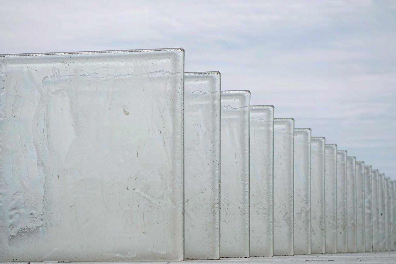 A long row of identical unlabeled frosted glass tiles standing upright on a pale concrete surface, receding into soft focus