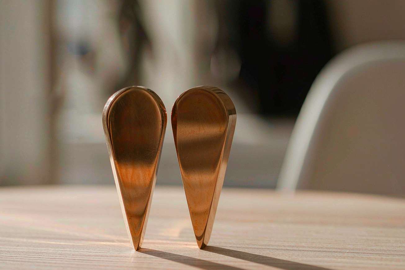 Two small blank brass sound-wave tiles standing upright on a pale wood table, warm light raking across their smooth unmarked surfaces
