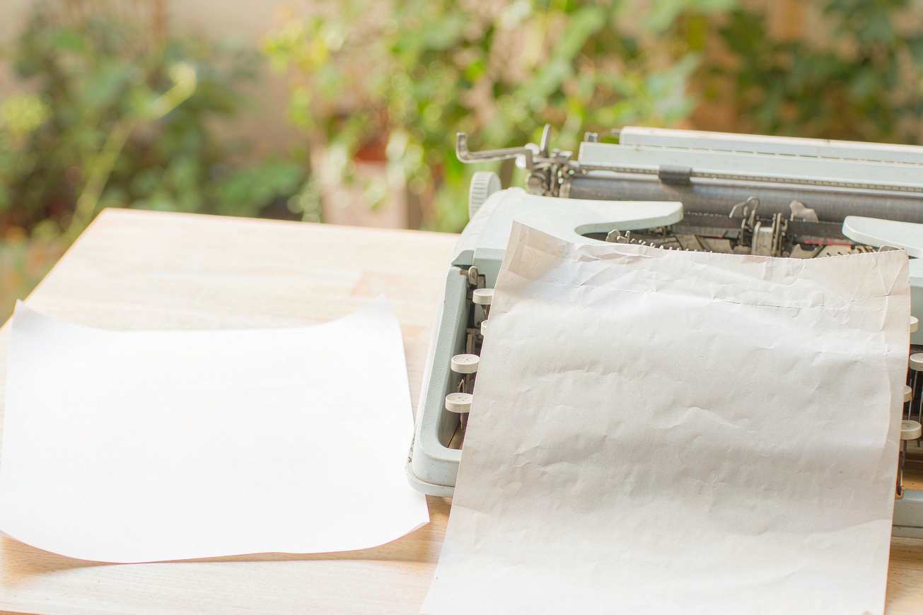 A blank sheet of paper emerging from a pale typewriter on a light wood desk, the page entirely unprinted