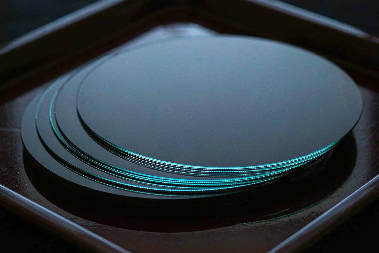 A cross-section of a densely layered silicon wafer stack on a dark anodized tray, thin cyan light catching the edges of each layer